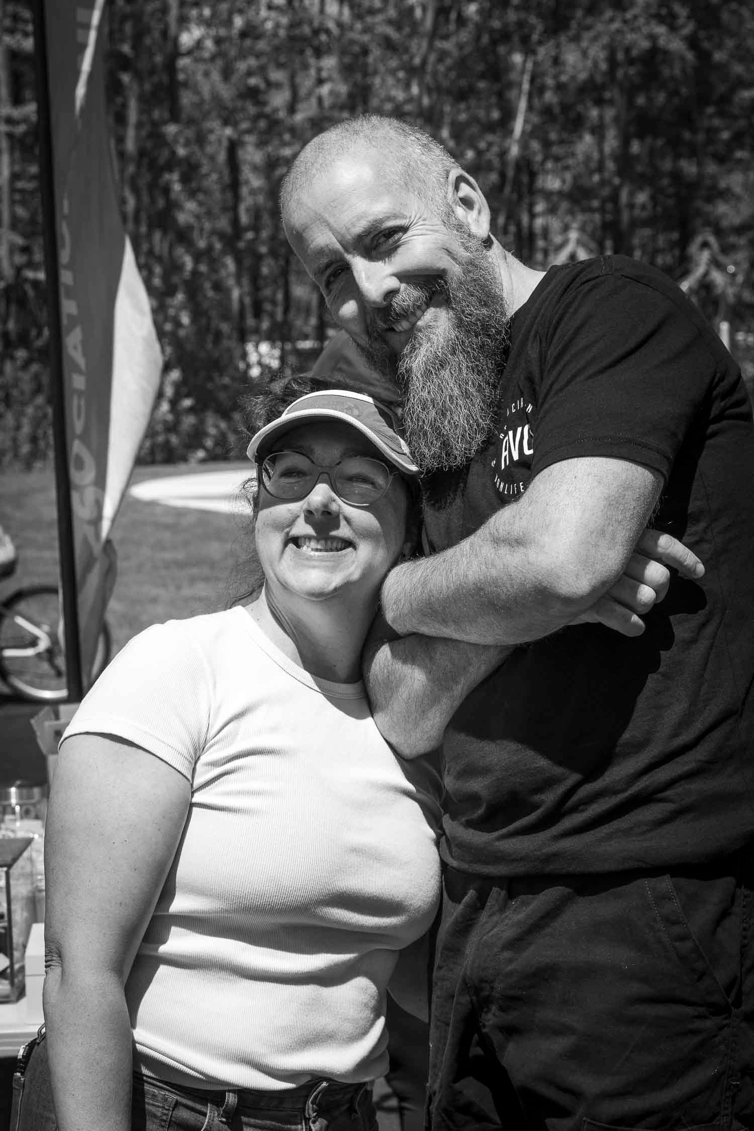 Black and white photo of a smiling couple outdoors, bearded bald man hugging a woman in a cap and white t-shirt.