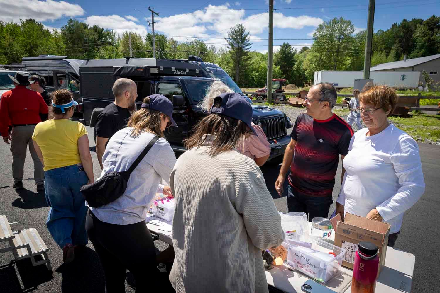 Group of people gathering outdoors around a table with food and supplies on a sunny day near parked vehicles.