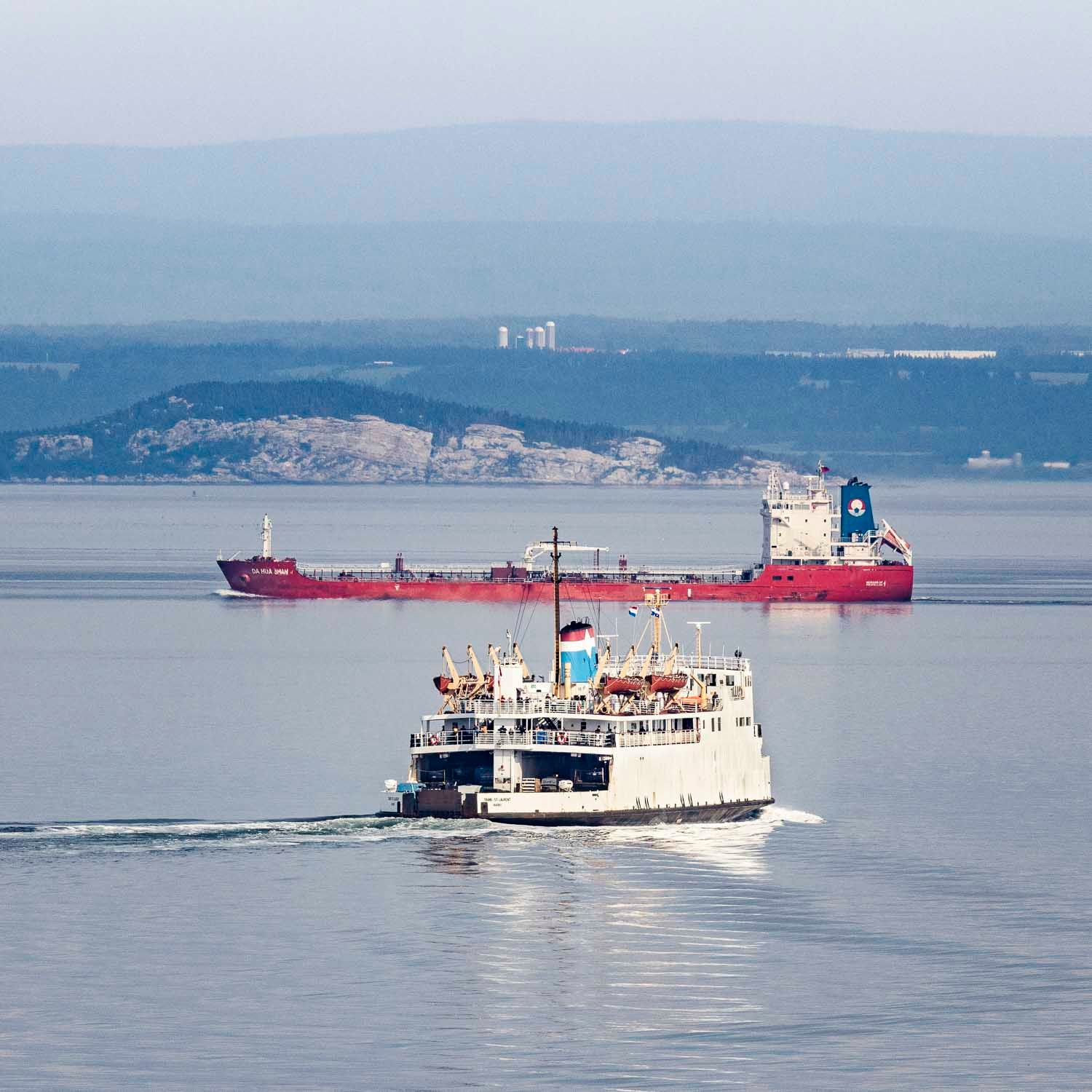 Traversier Saint-Siméon vers R-d-L.
A white ferry and red cargo ship sail on calm blue waters with a hilly coastline in the background.