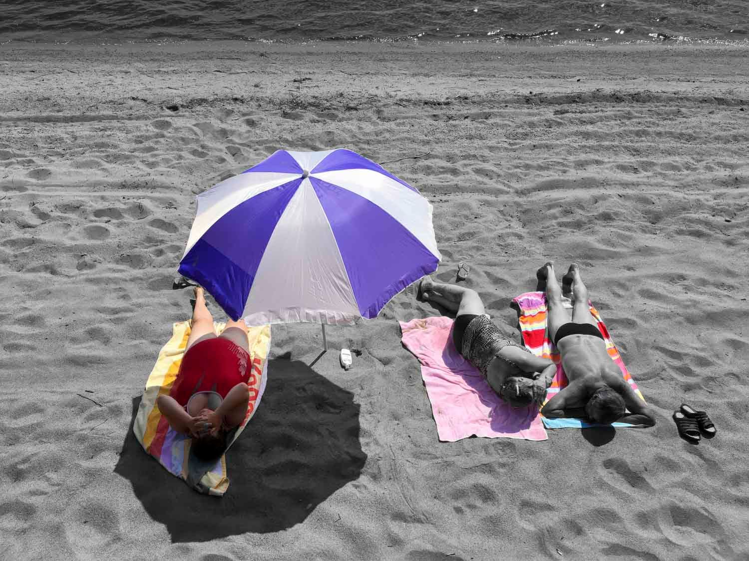 Two people sunbathing on a beach under a blue and white umbrella on colorful towels in a selective color photo.