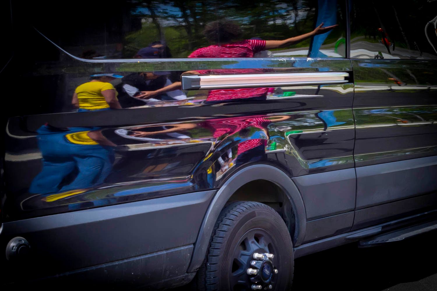 Colorful reflections of people visible on the shiny dark surface of a parked vehicle.
