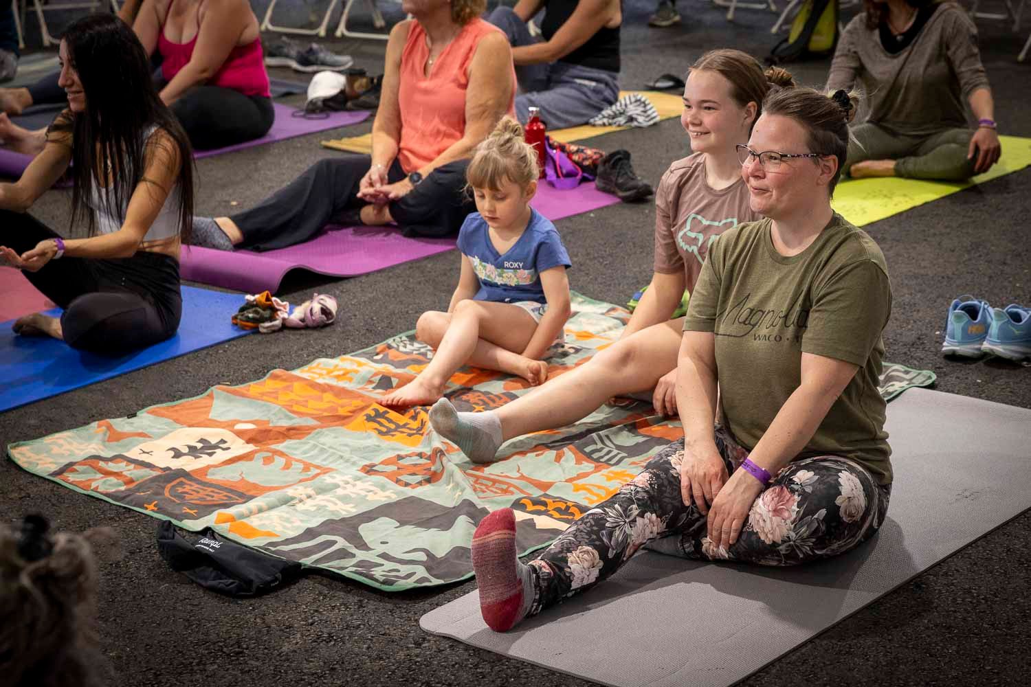 Adults and a child sit cross-legged on colorful yoga mats during a family yoga class indoors.