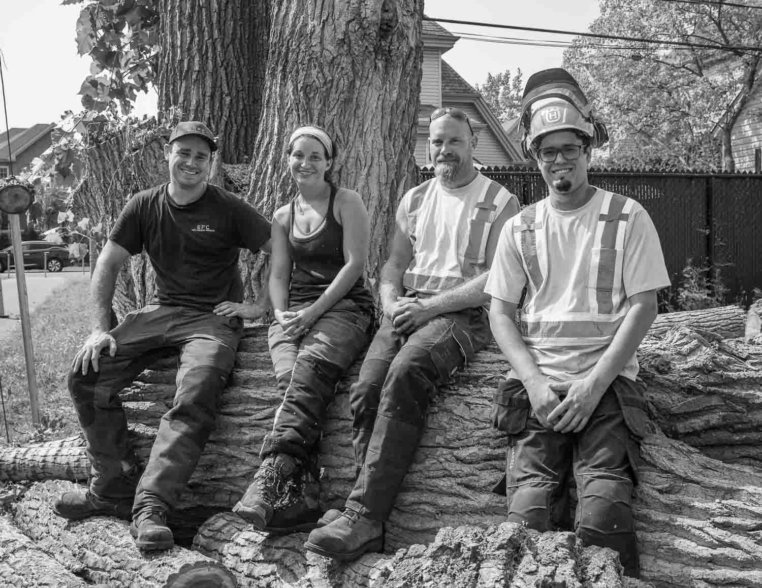 Four tree service crew members sitting on large cut logs beside a tree in black and white.