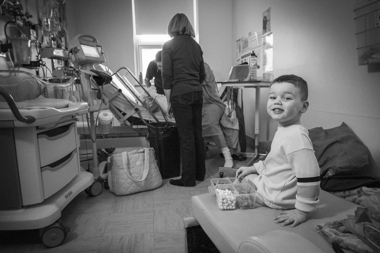 Black and white photo of smiling boy sitting in hospital room while adult tends to medical equipment.