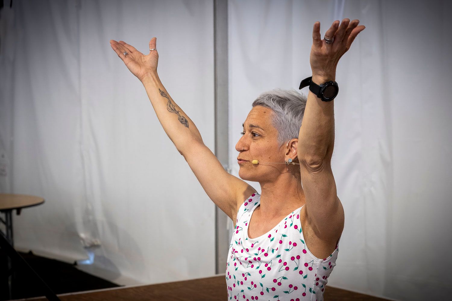 Woman with short gray hair raises arms overhead during yoga or dance class inside white tent.
