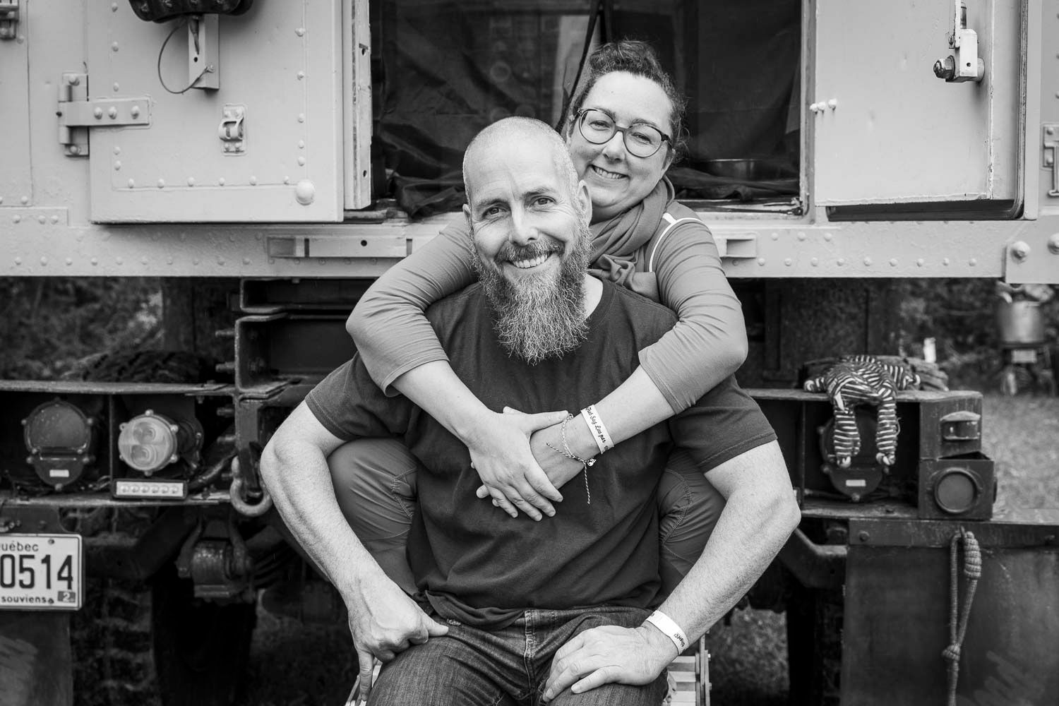 Black and white close-up portrait of a bearded bald man and smiling woman embracing in front of a vehicle.