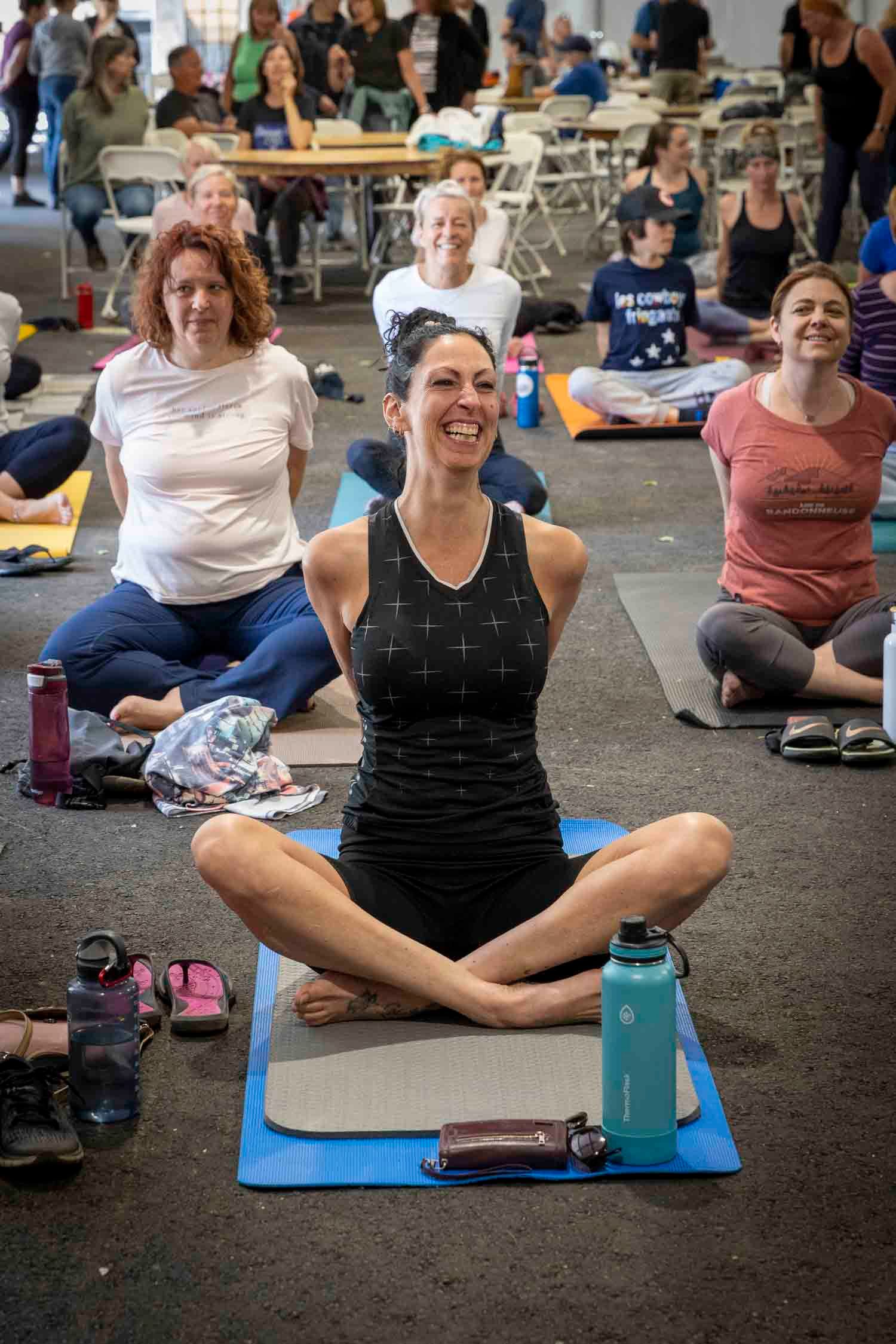 Woman smiling in lotus pose on blue yoga mat during group yoga class indoors.