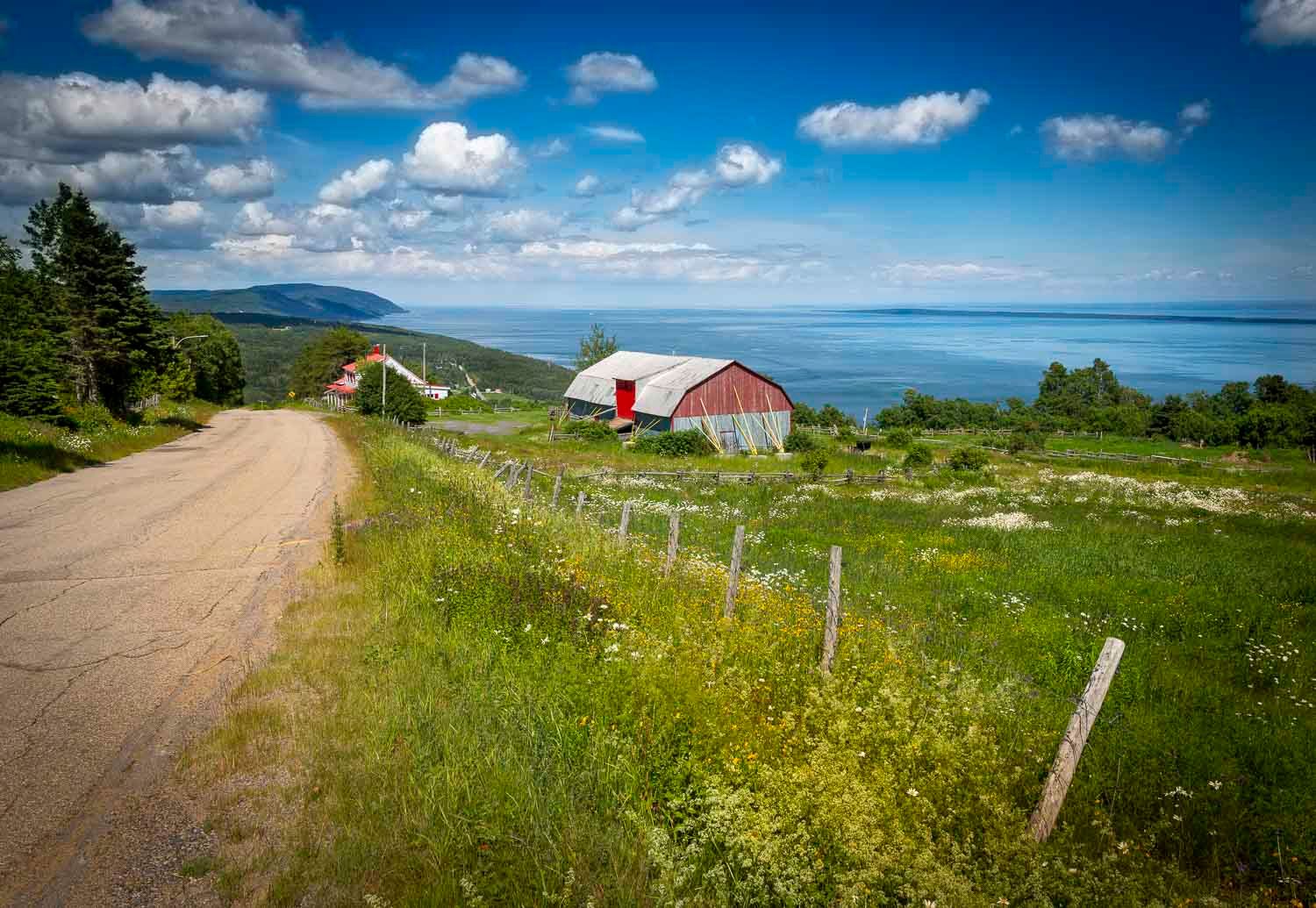 Rural country road with red barn, green meadows, and scenic ocean view under blue sky.