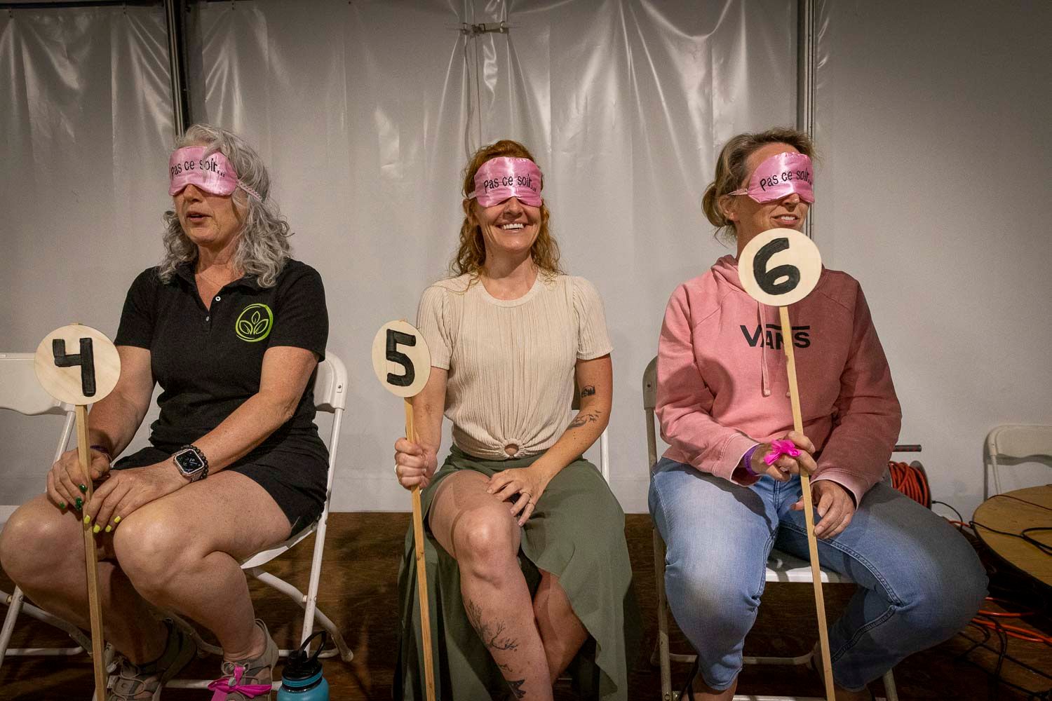 Three blindfolded women holding numbered paddles sitting on chairs at a fun tasting competition event.