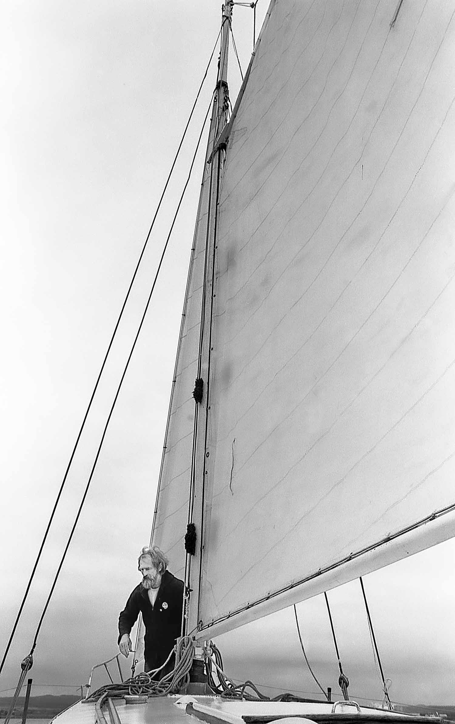 Black and white photo of a sailor on the bow of a sailboat with a large mainsail billowing against an overcast sky.