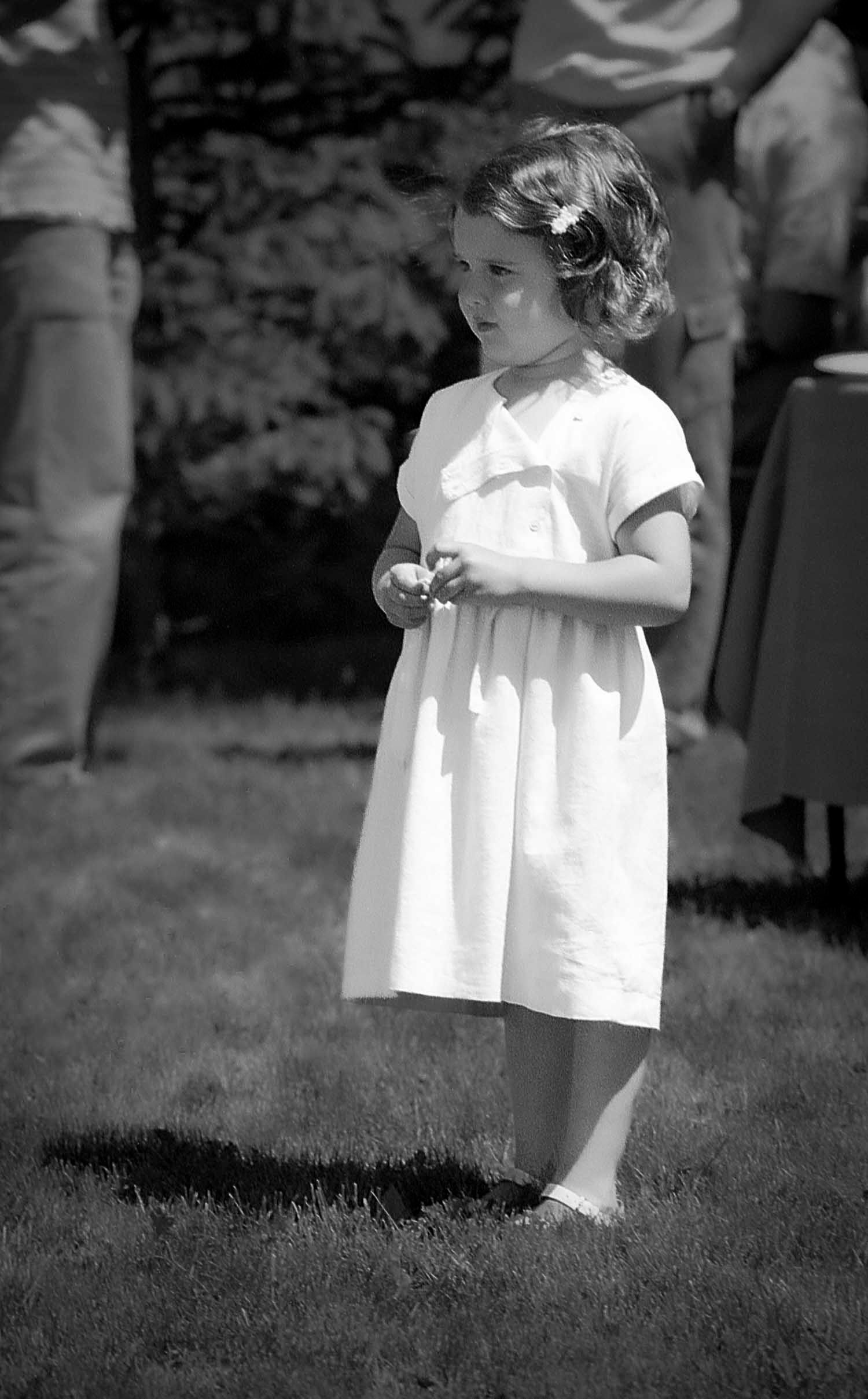Young girl in a white dress with a flower in her hair standing on grass in a black and white photo.