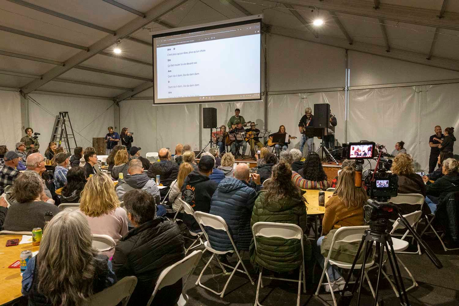 Audience seated at tables facing a presenter and projection screen inside a large event tent.