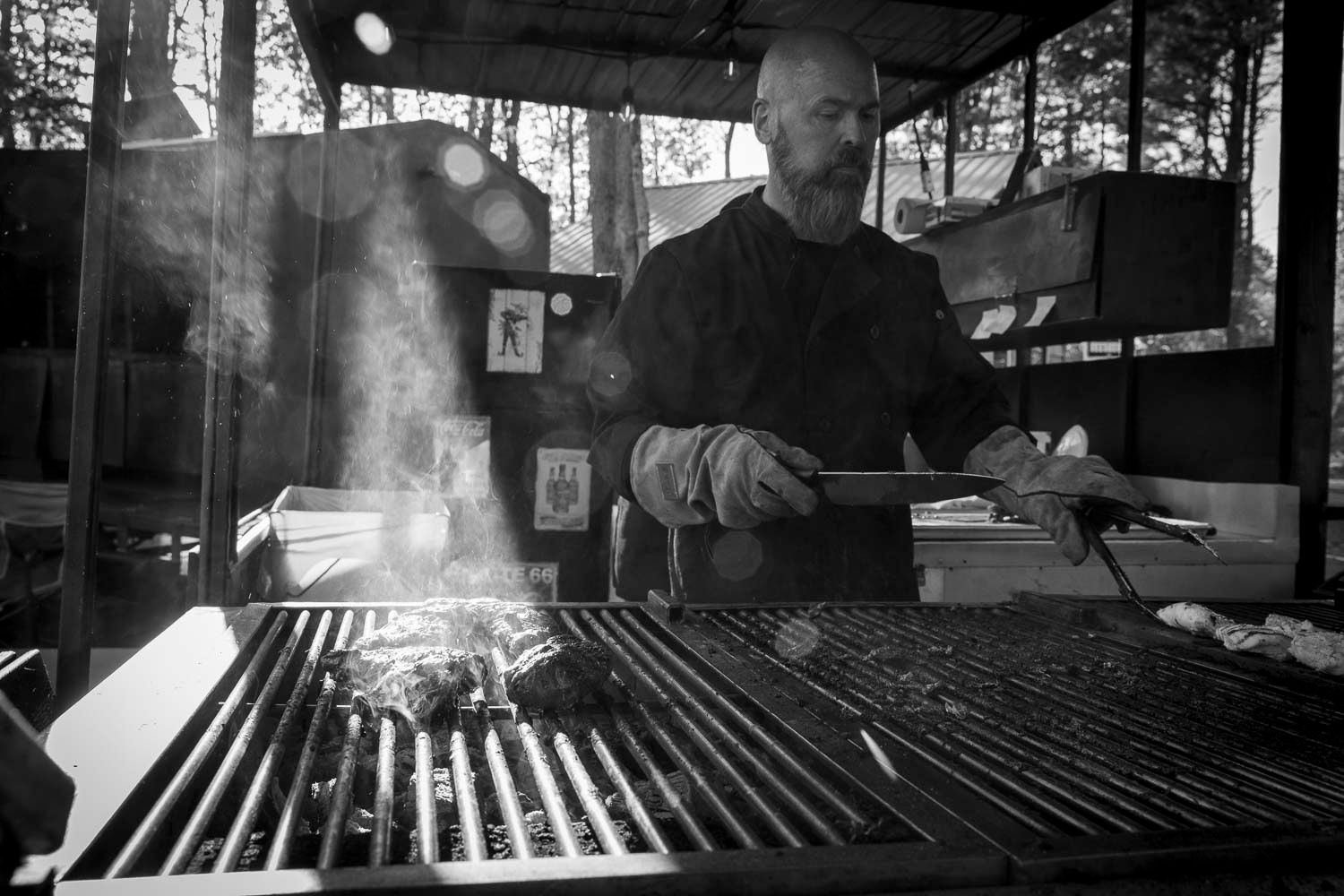 Chef grilling meat over open flames in a rustic outdoor kitchen, black and white photo.
