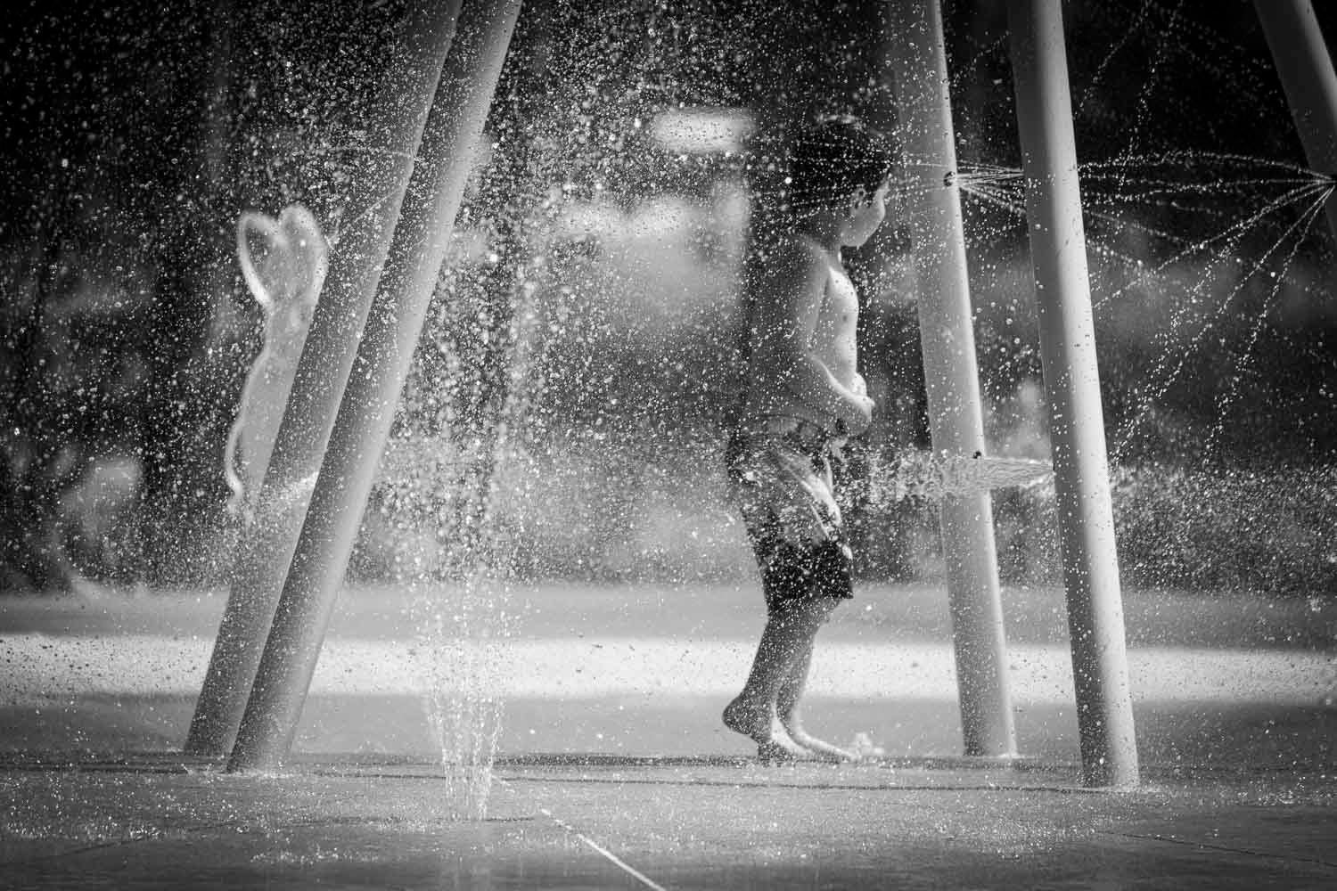 Black and white photo of child playing in outdoor splash pad water fountain.