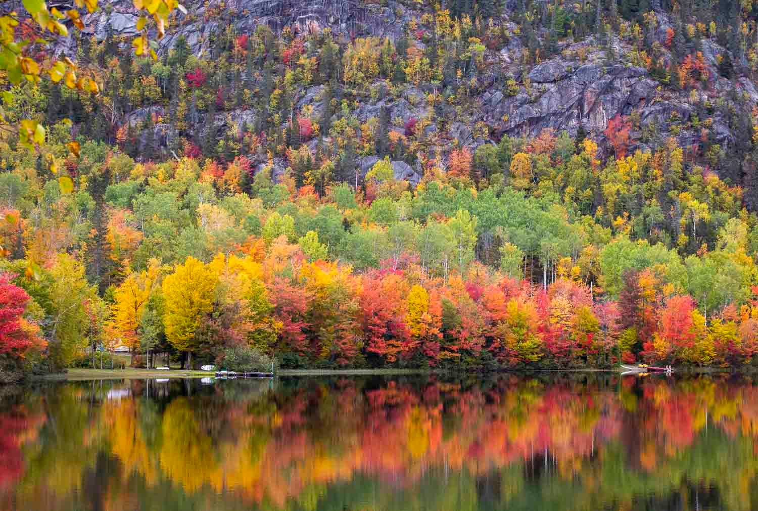 Paysage automne.
Vibrant autumn foliage reflecting on a calm lake with colorful red, orange, and yellow trees against a rocky hillside.
