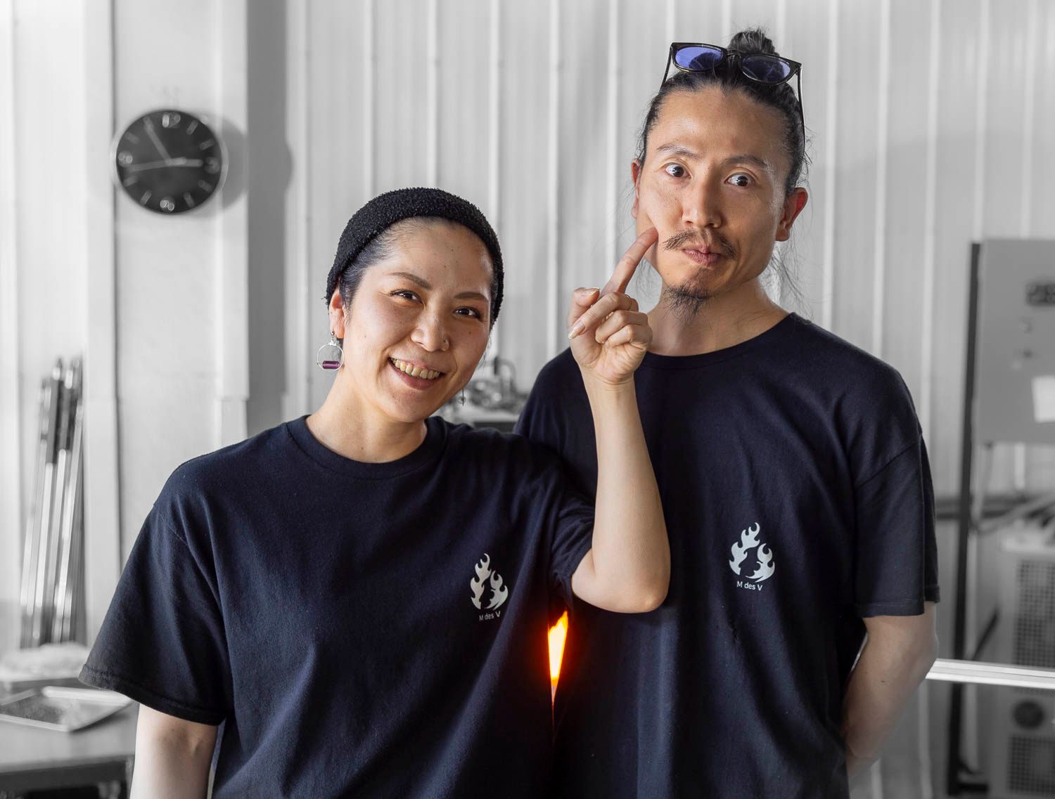 Two smiling artists in matching navy blue t-shirts posing together in a glassblowing studio.