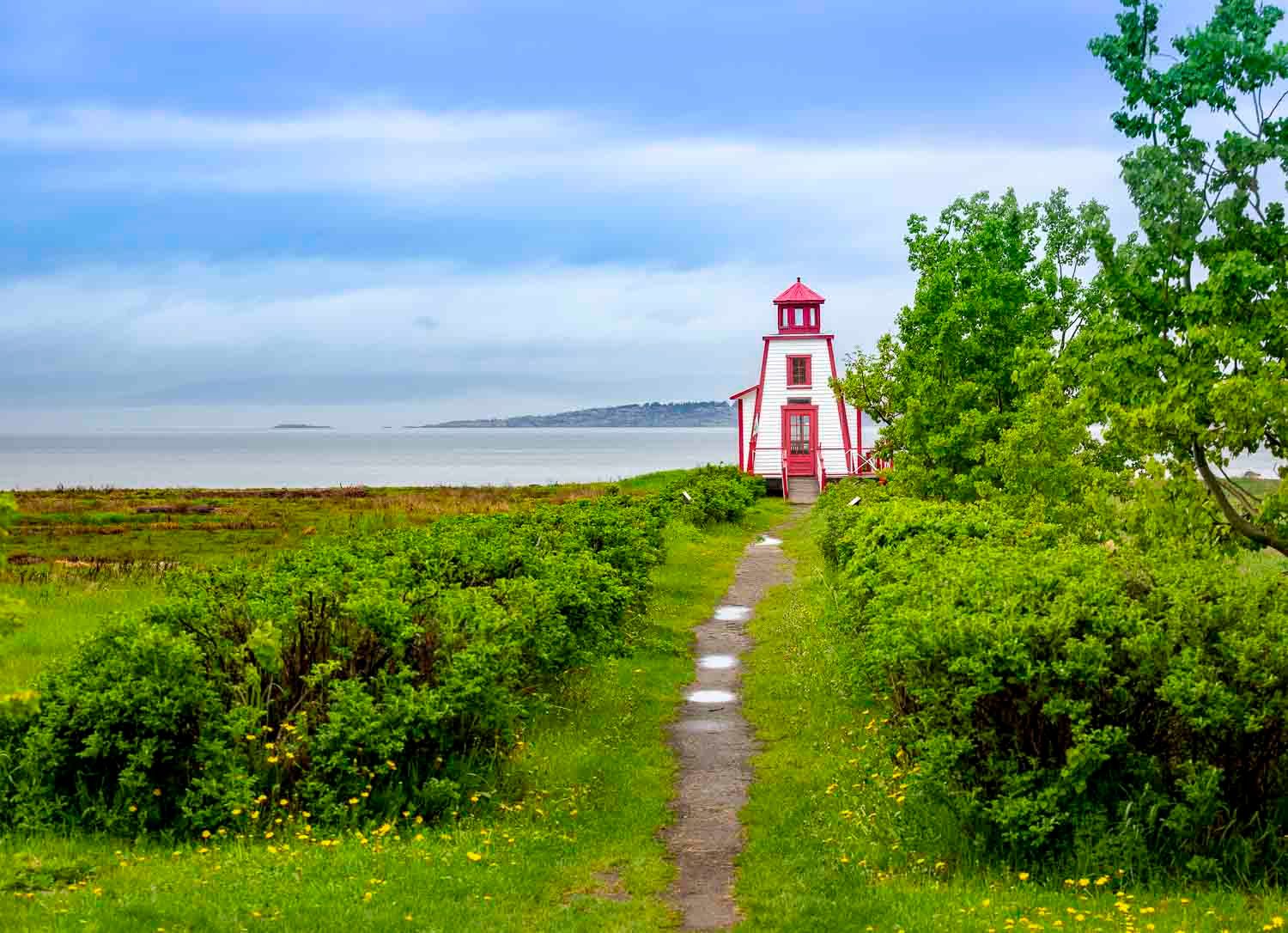 Red and white lighthouse along a green coastal path with ocean views under cloudy skies.