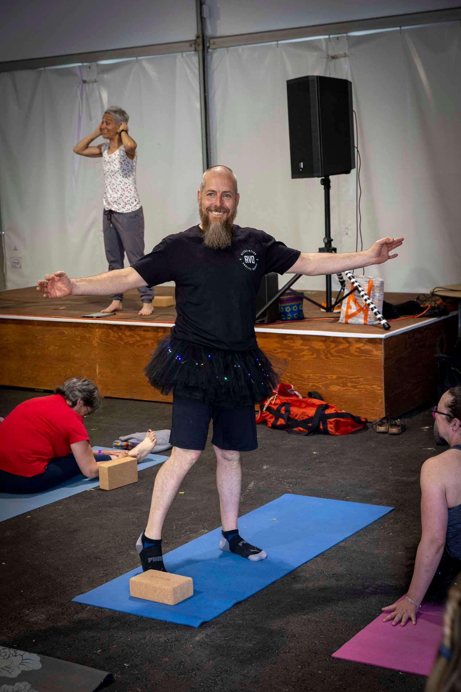 Bald bearded man wearing tutu balances on yoga block with arms outstretched during indoor yoga class.
