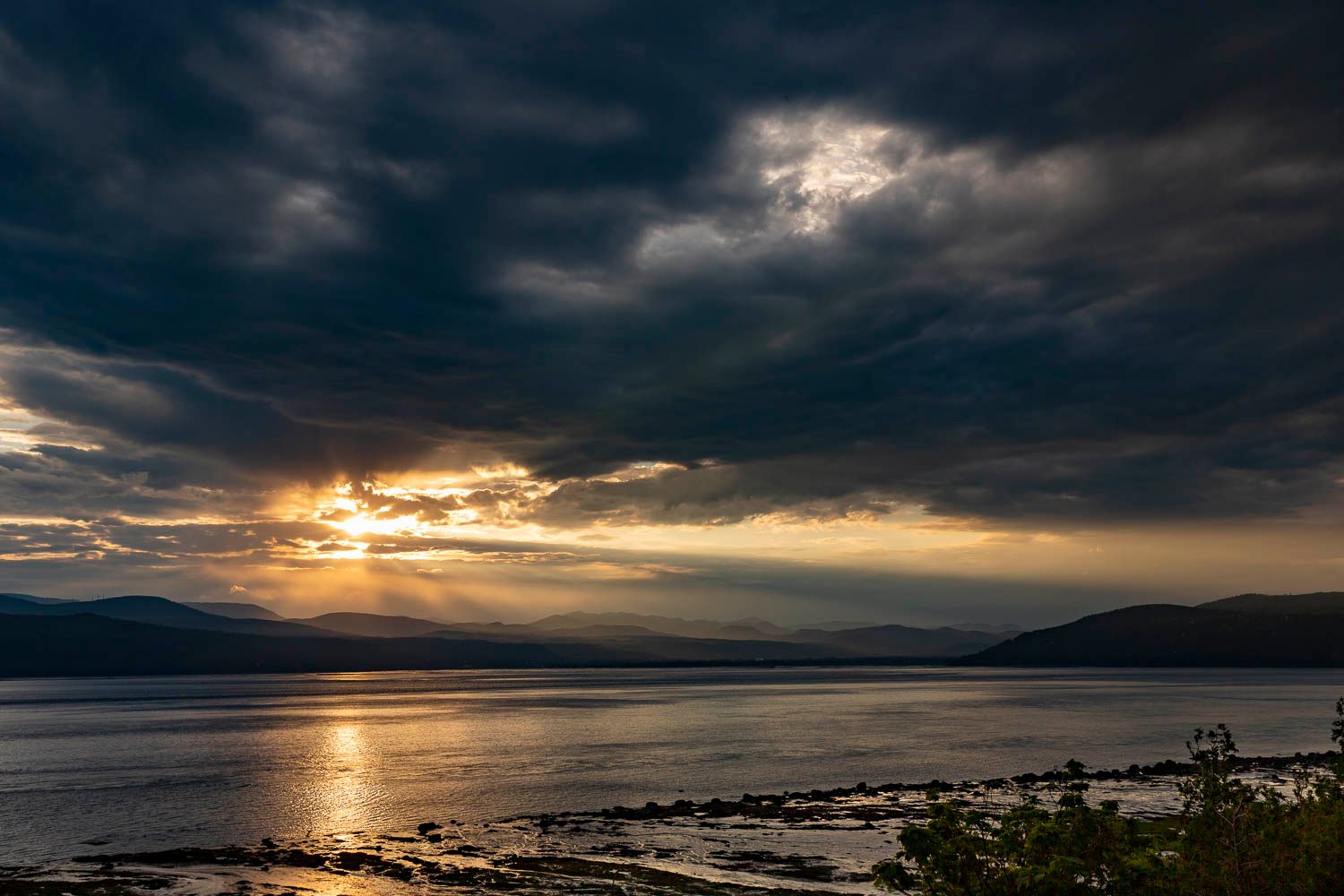 Dramatic dark storm clouds with sunbeams breaking through over a calm coastal inlet at sunset.
