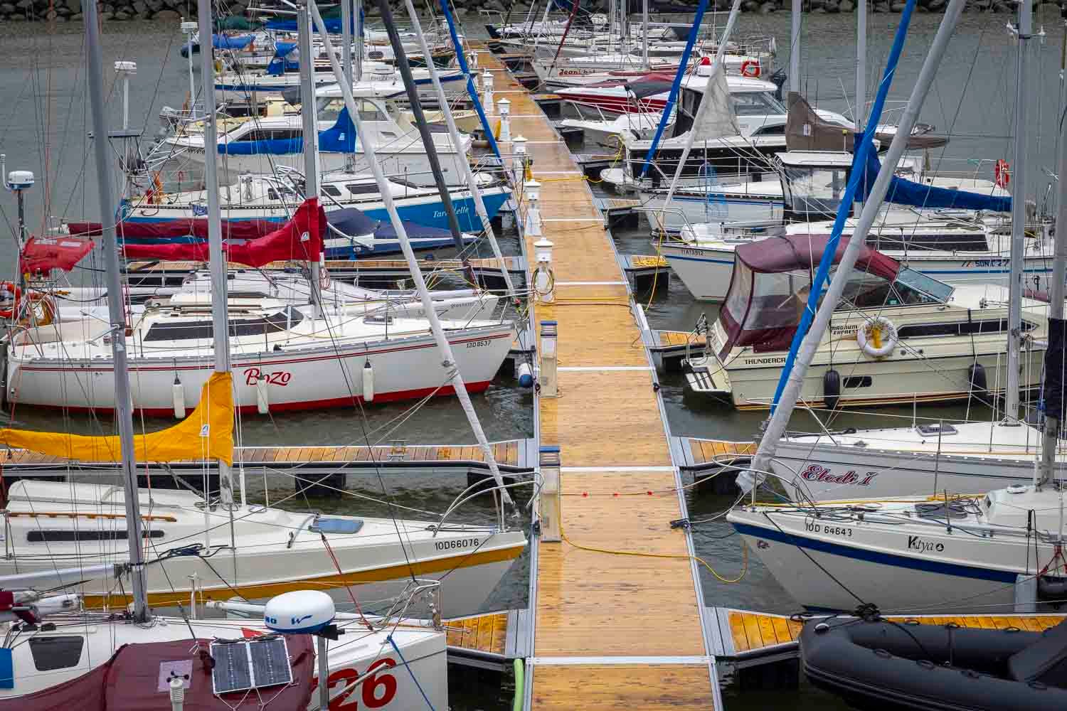 Aerial view of numerous sailboats docked along a wooden pier at a busy marina on an overcast day.