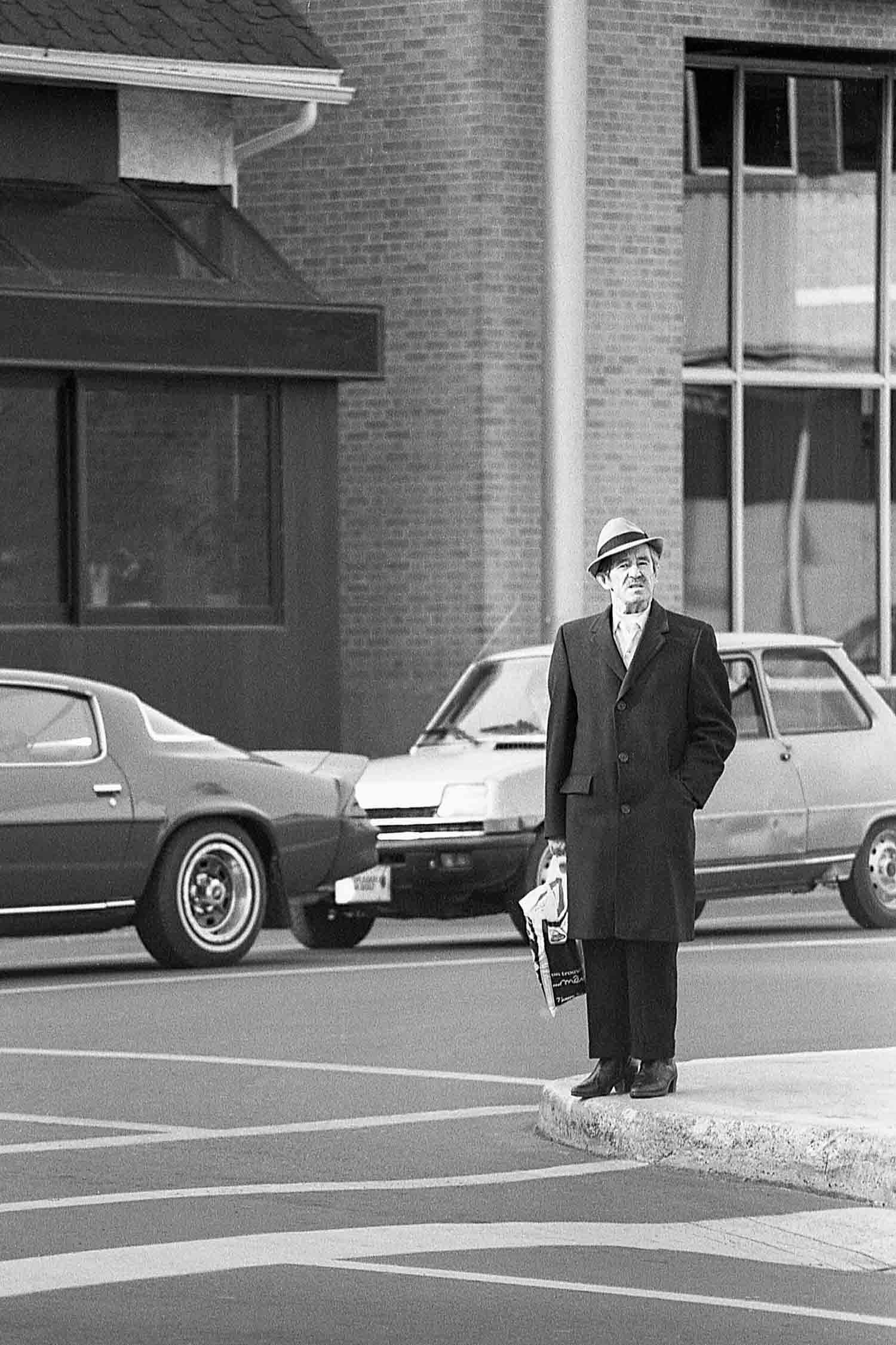 Elderly man in a dark coat and hat standing on a street corner holding a bag in a black and white photo.