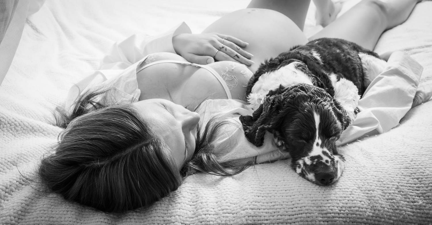Black and white photo of a pregnant woman lying on a bed with a black and white spaniel dog cuddled beside her.