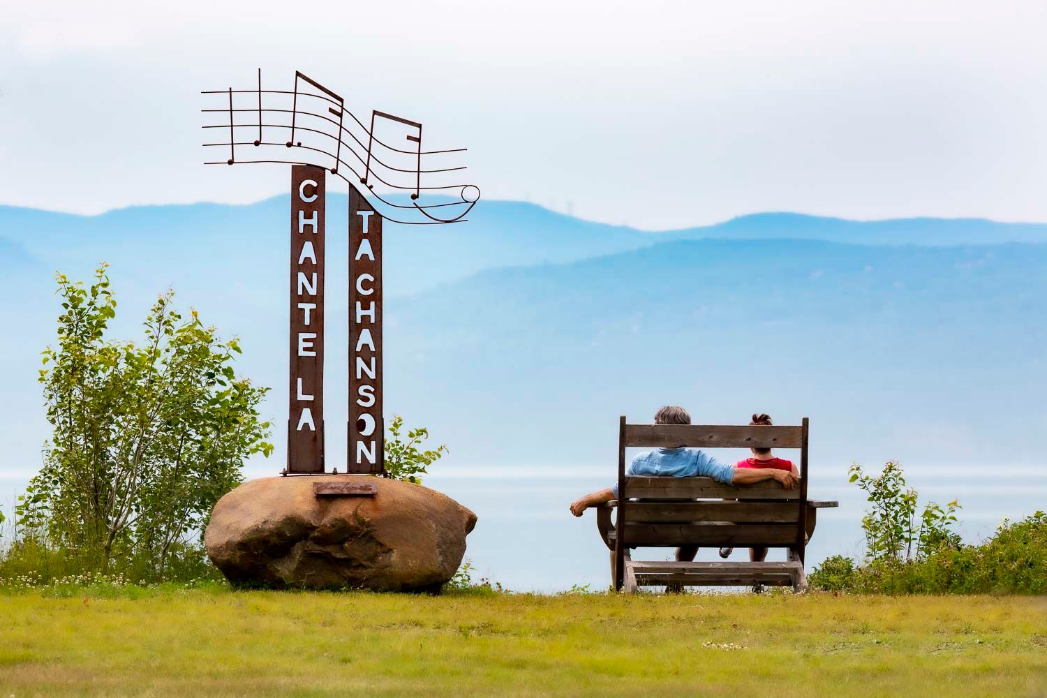 Havre musical de l'isle aux coudres
Two people sit on a bench overlooking misty mountains beside a  musical sign on a hilltop.