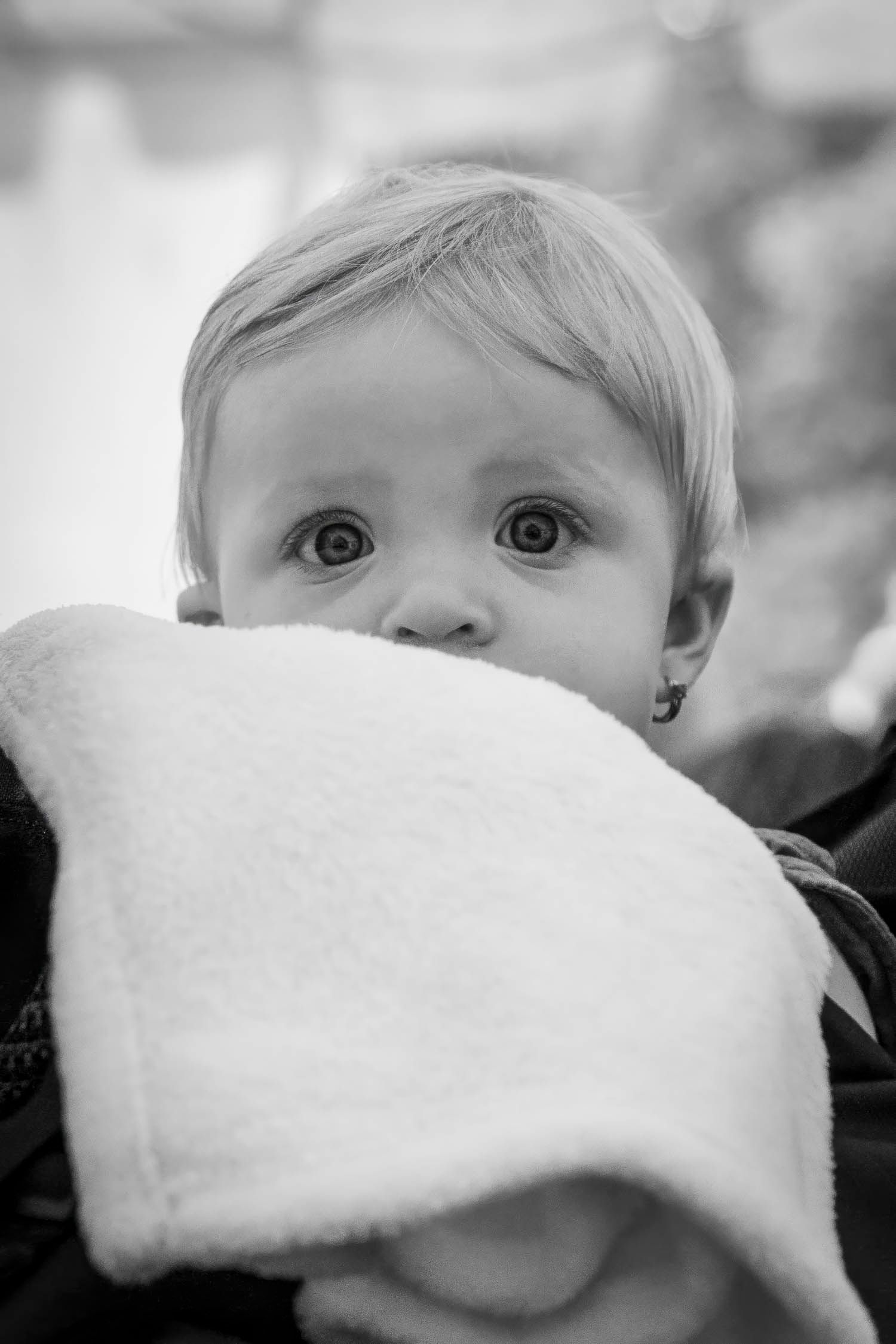 Black and white portrait of a curious baby peeking over a soft white blanket with wide expressive eyes.