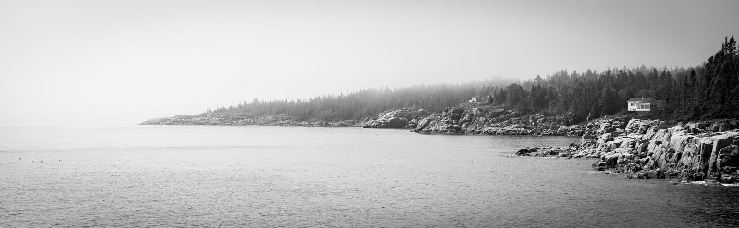 Les Escoumains
Black and white panoramic coastal landscape with rocky shoreline, dense pine forest, and a small house in misty fog.
