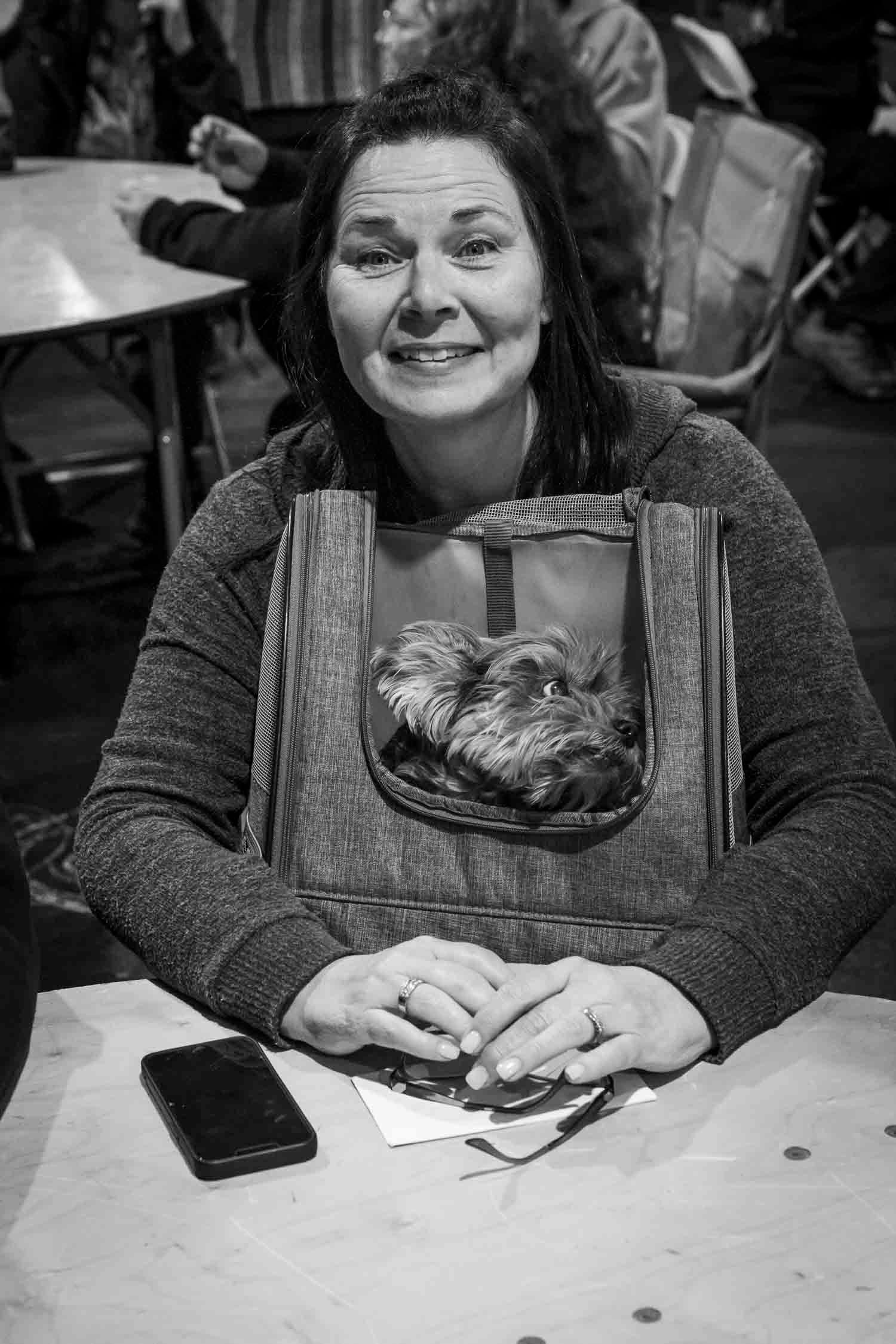 Smiling woman sitting at a table with a small dog in a carrier bag in black and white photo.