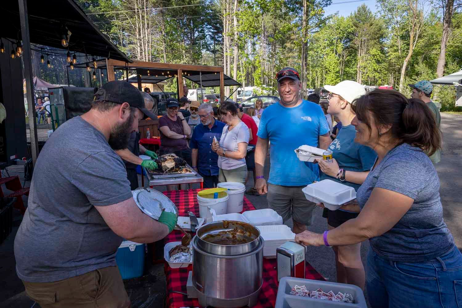 People serving and receiving food at an outdoor community cookout event near a wooded area.