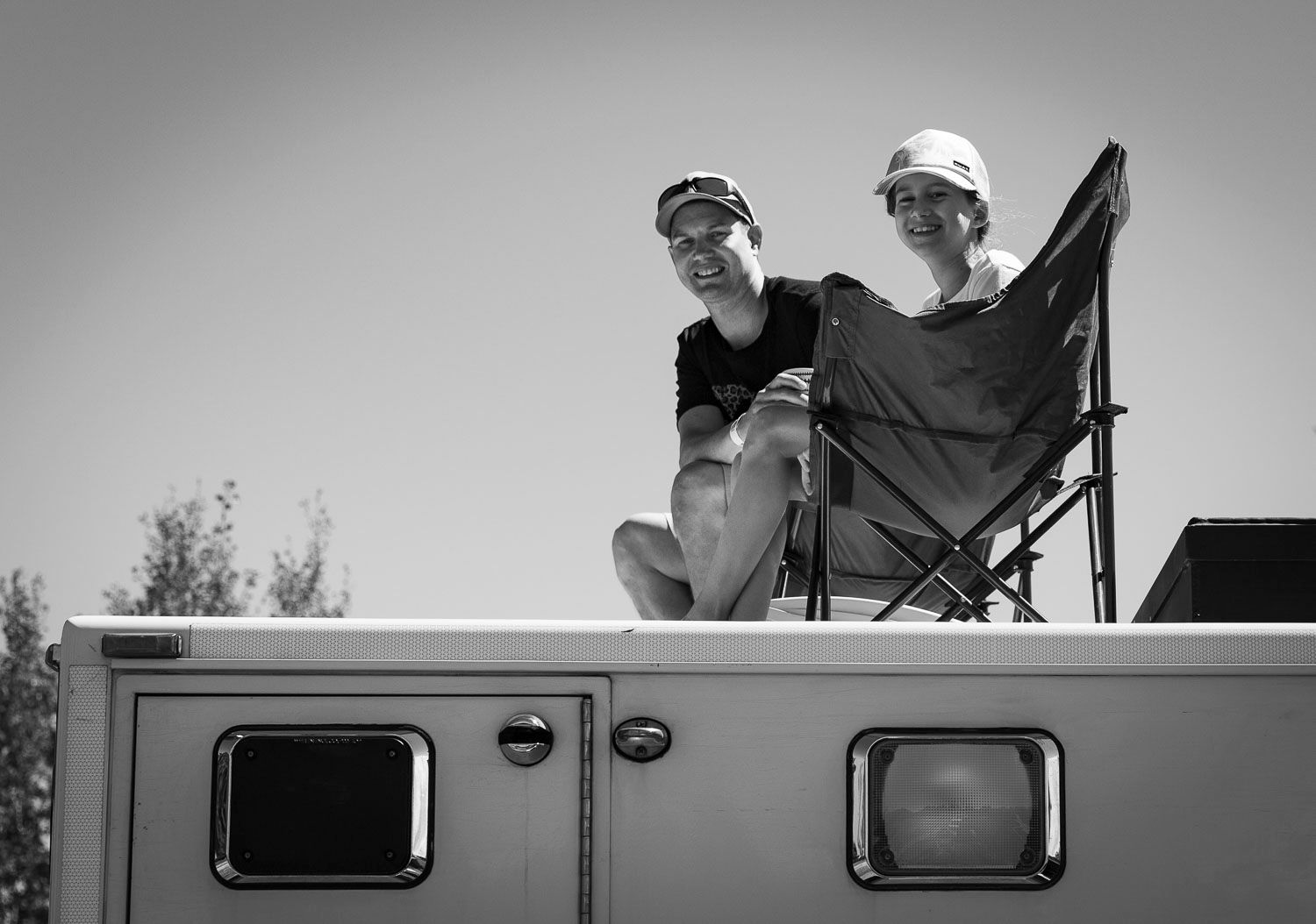 Two people sitting on top of an RV roof with a folding chair on a sunny day in black and white.