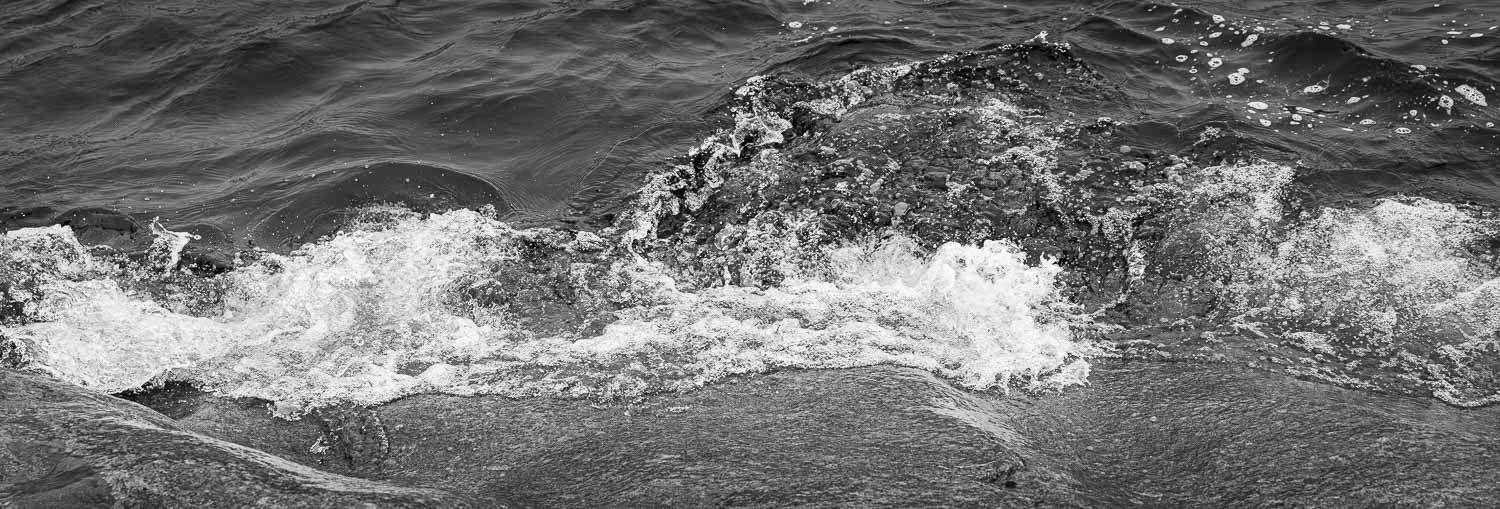 Black and white panoramic photo of ocean waves crashing over rocks along the shoreline.