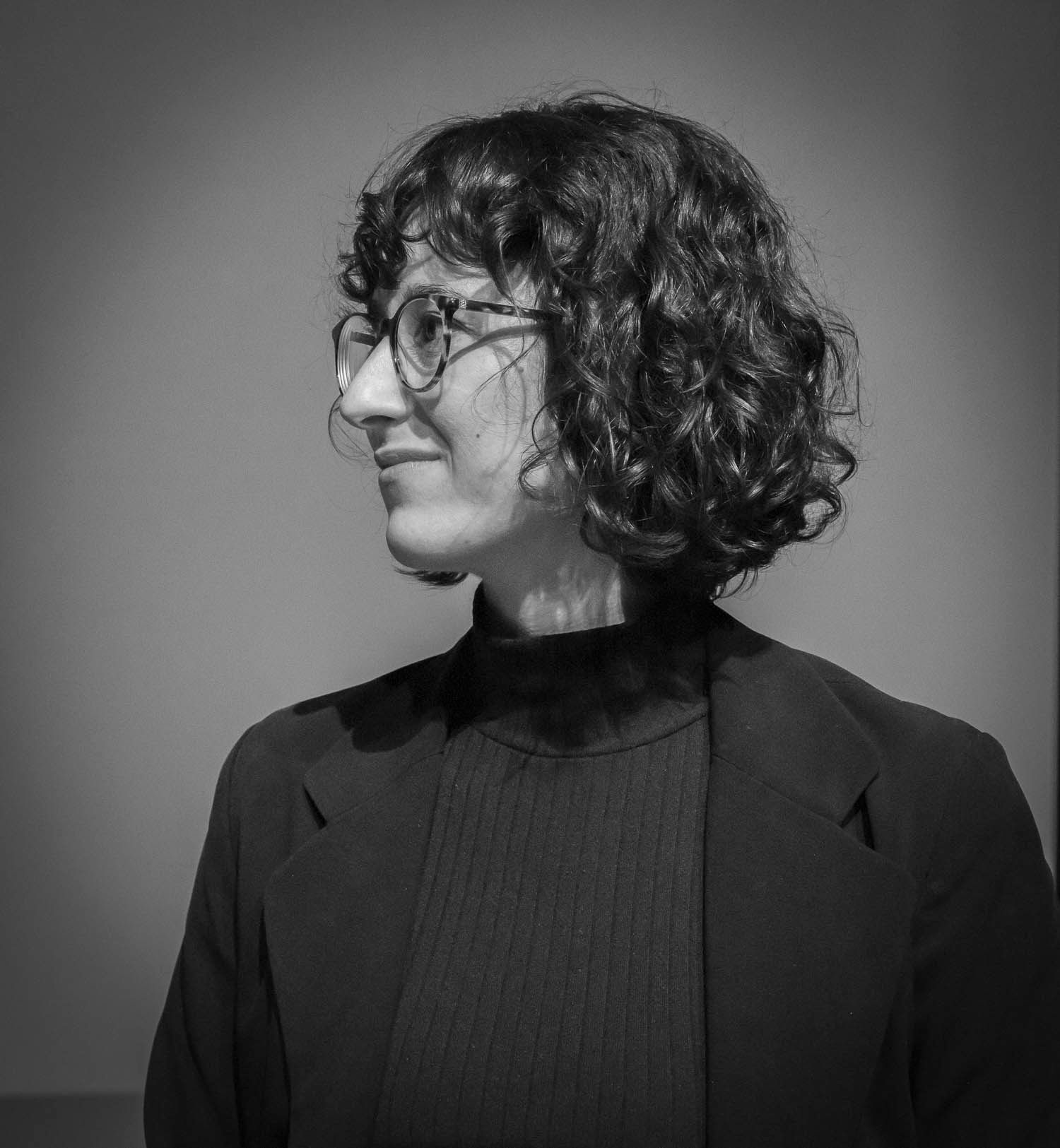 Black and white portrait of a person with curly hair and glasses wearing a turtleneck, looking sideways.