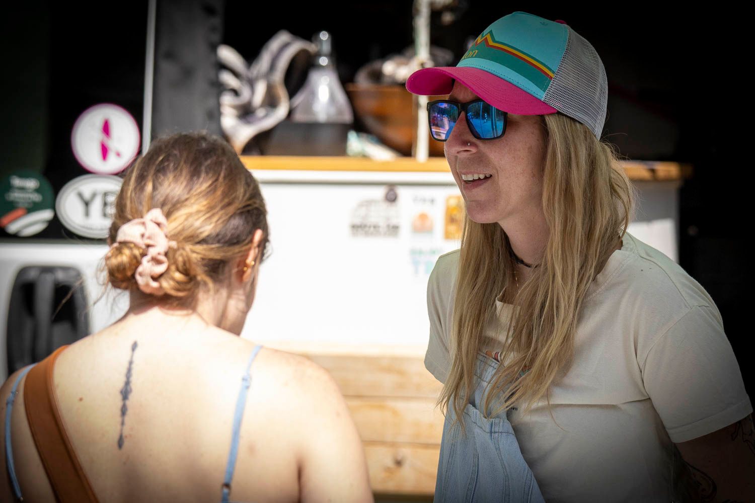 Two women conversing at an outdoor food stand, one wearing colorful sunglasses and a cap.