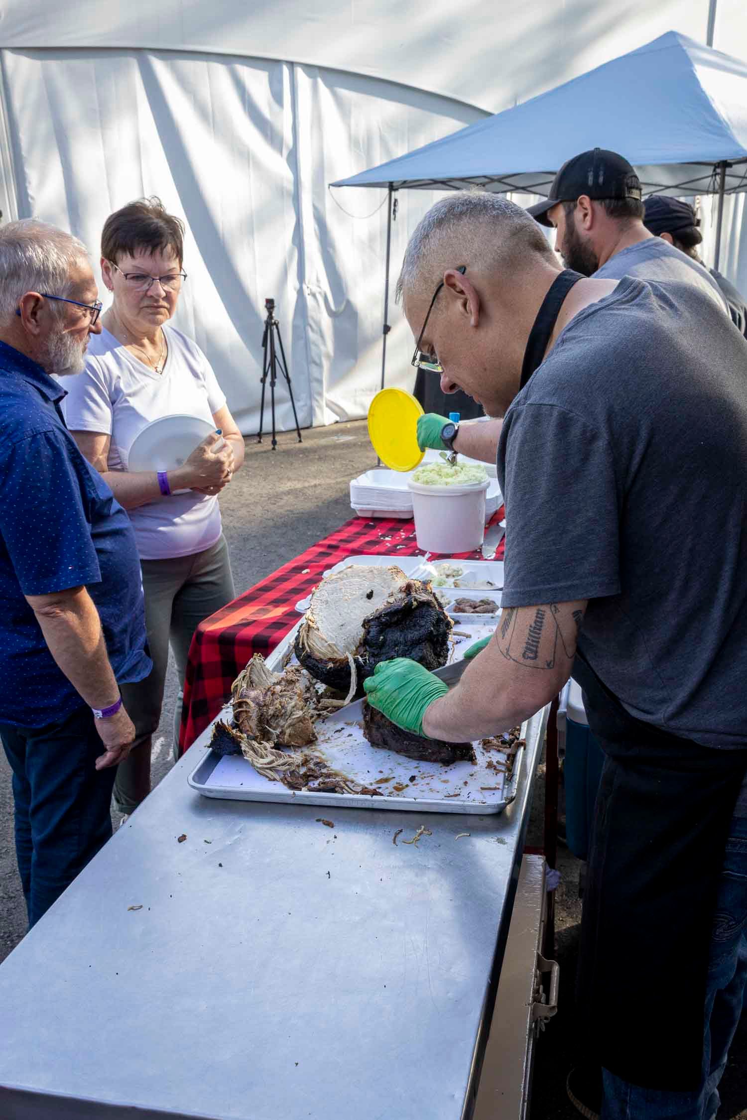 Man with tattoos cuts large chocolate cake at outdoor food festival while guests watch under a white tent.