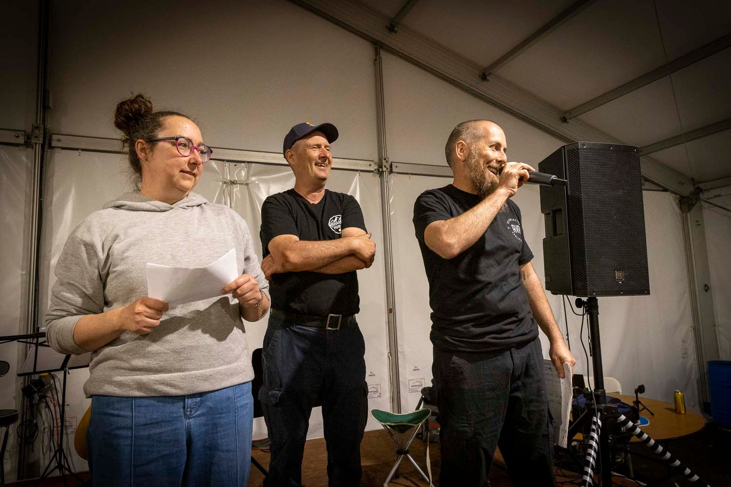 Three people on stage at an outdoor event, one speaking into a microphone under a white tent.