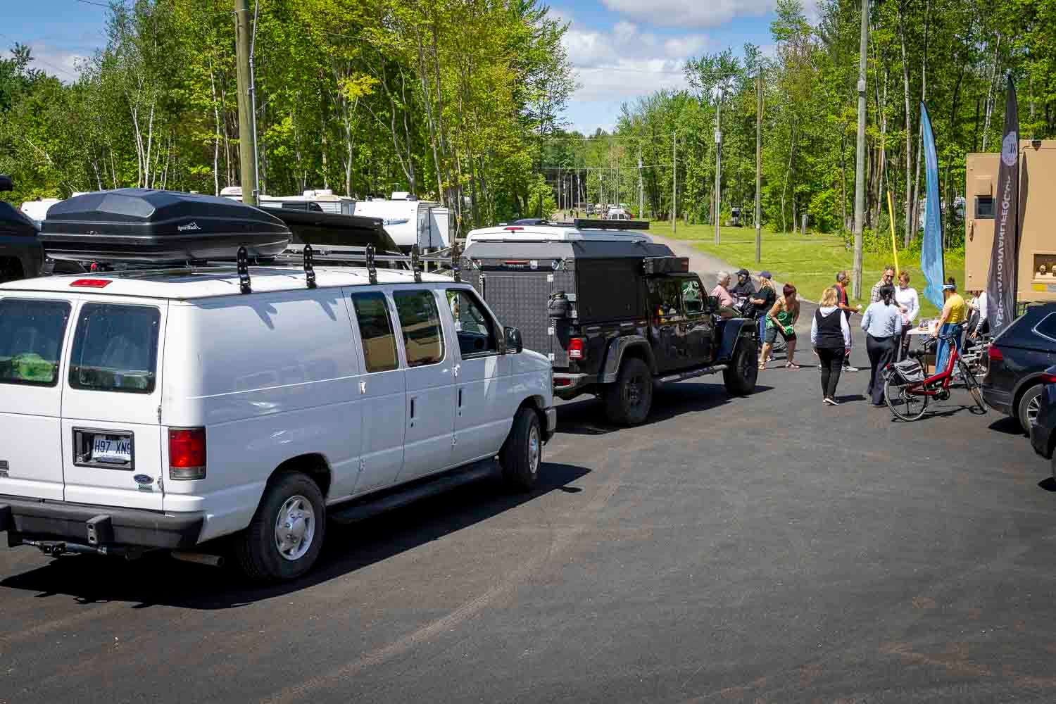 White van and black truck with camper trailer parked in sunny lot with people gathered nearby, surrounded by green trees.
