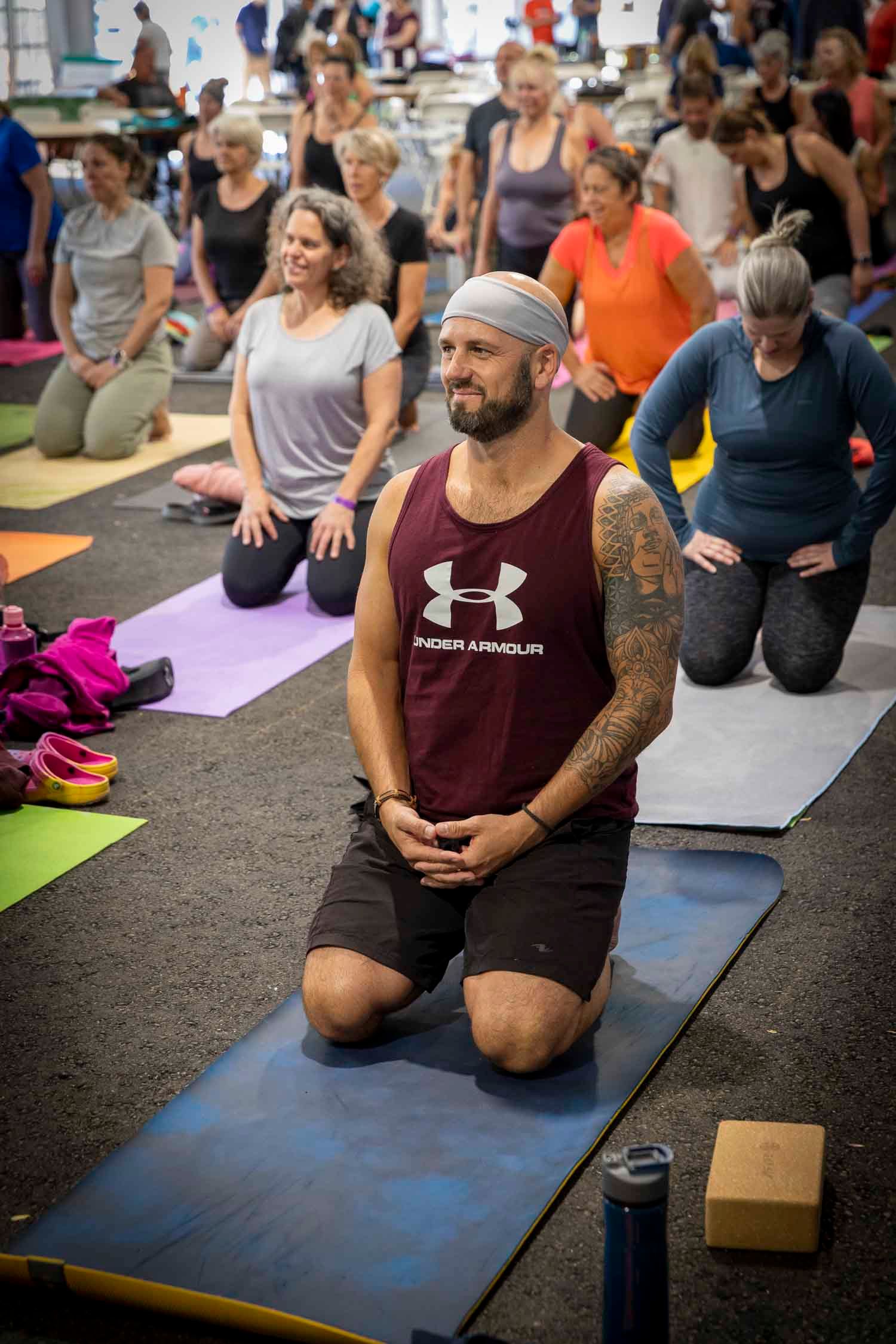 Man in Under Armour tank top kneeling on yoga mat leading a large group yoga class indoors.