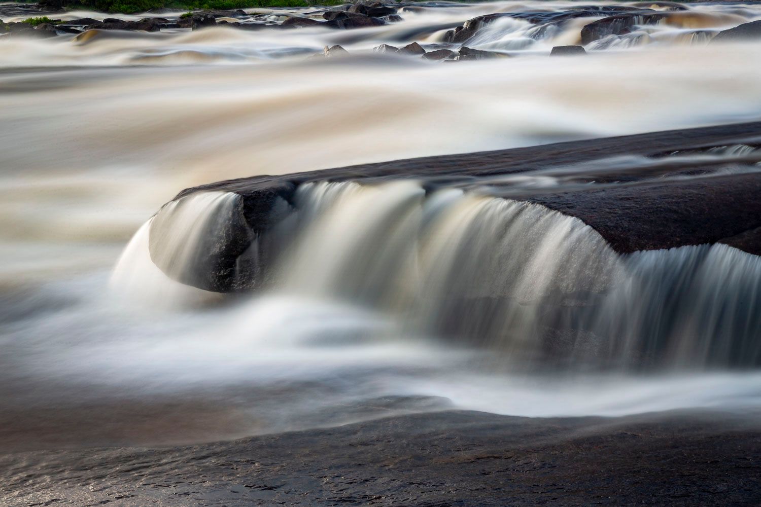 Long exposure color photograph of smooth silky waterfall flowing around a large boulder on rocky riverbed.
