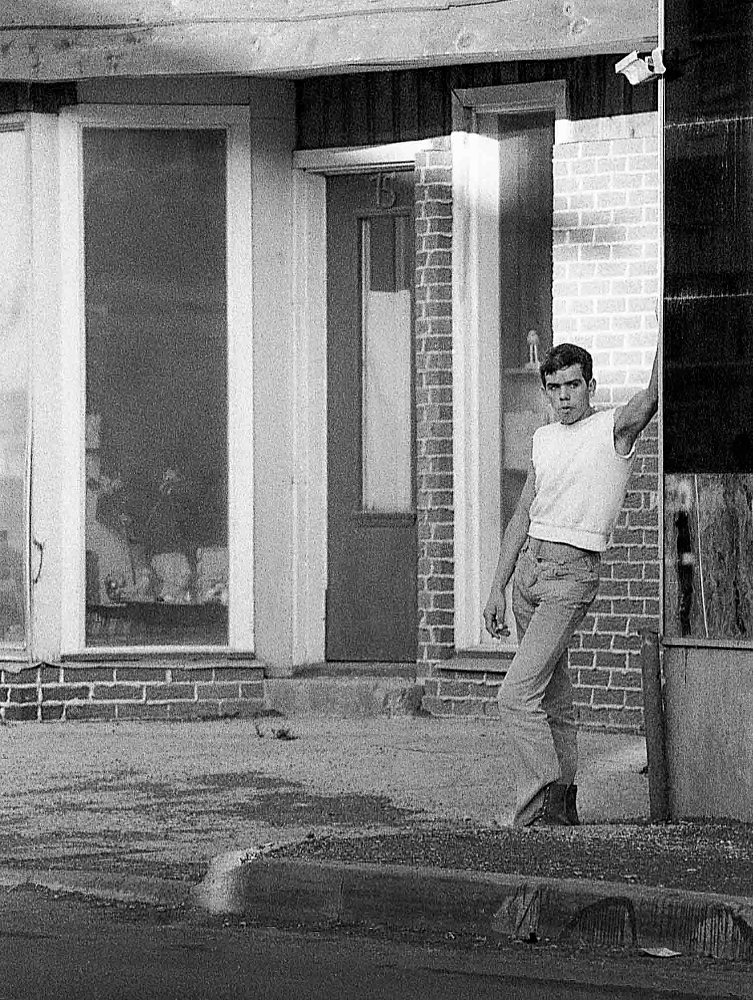 Young man in white t-shirt and jeans leaning against a brick building in a black and white street photograph.