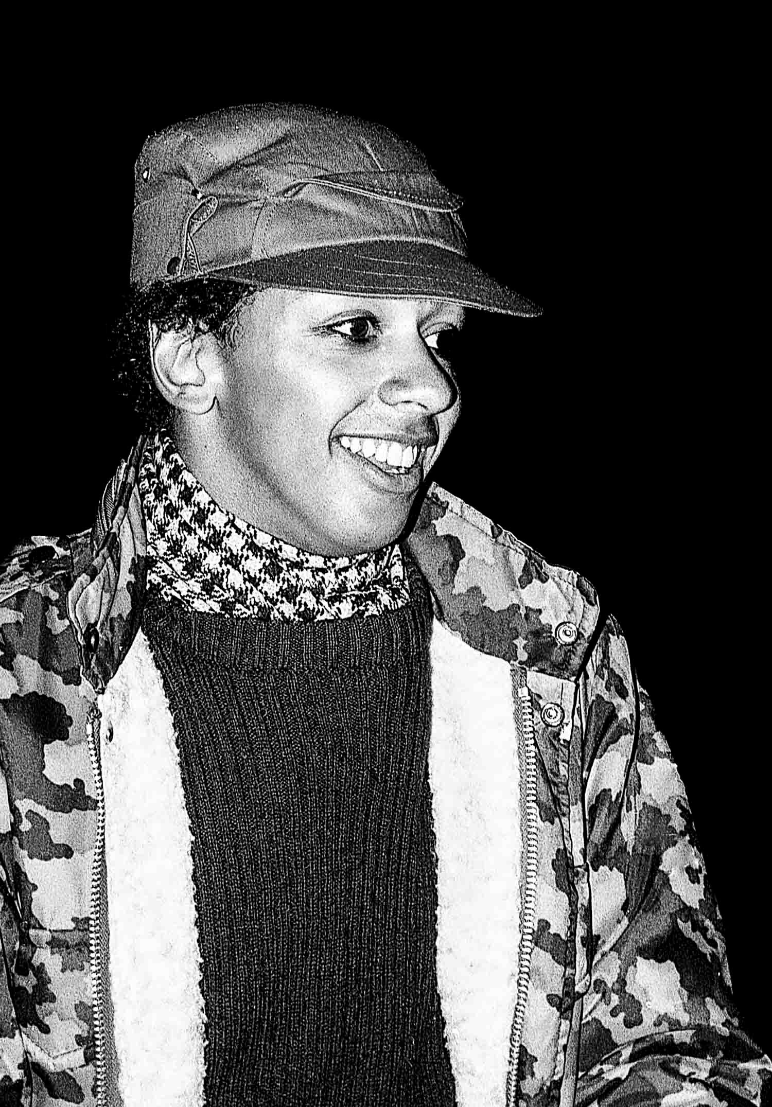 Black and white photo of a smiling person wearing a camo jacket, cap, and houndstooth scarf.