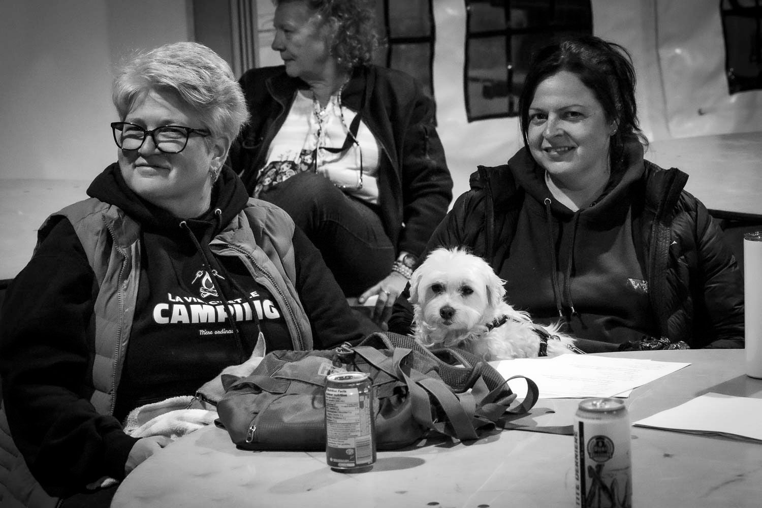 Two women smile at a table with a small white fluffy dog between them in a black and white photo.