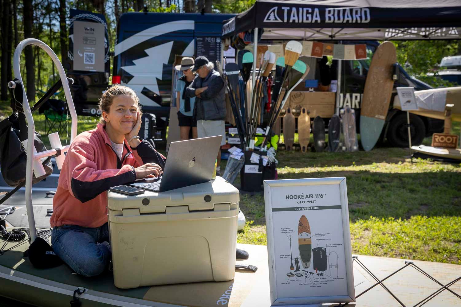 Woman working on laptop at outdoor vendor booth displaying Taiga Board paddleboards at an outdoor expo event.