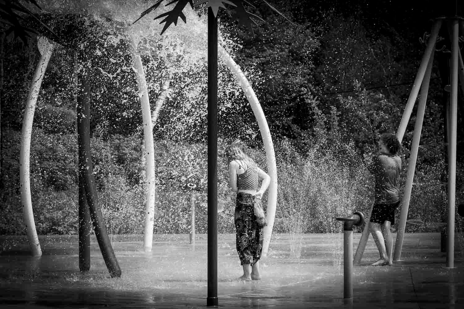 Black and white photo of two people standing in outdoor water splash pad area.