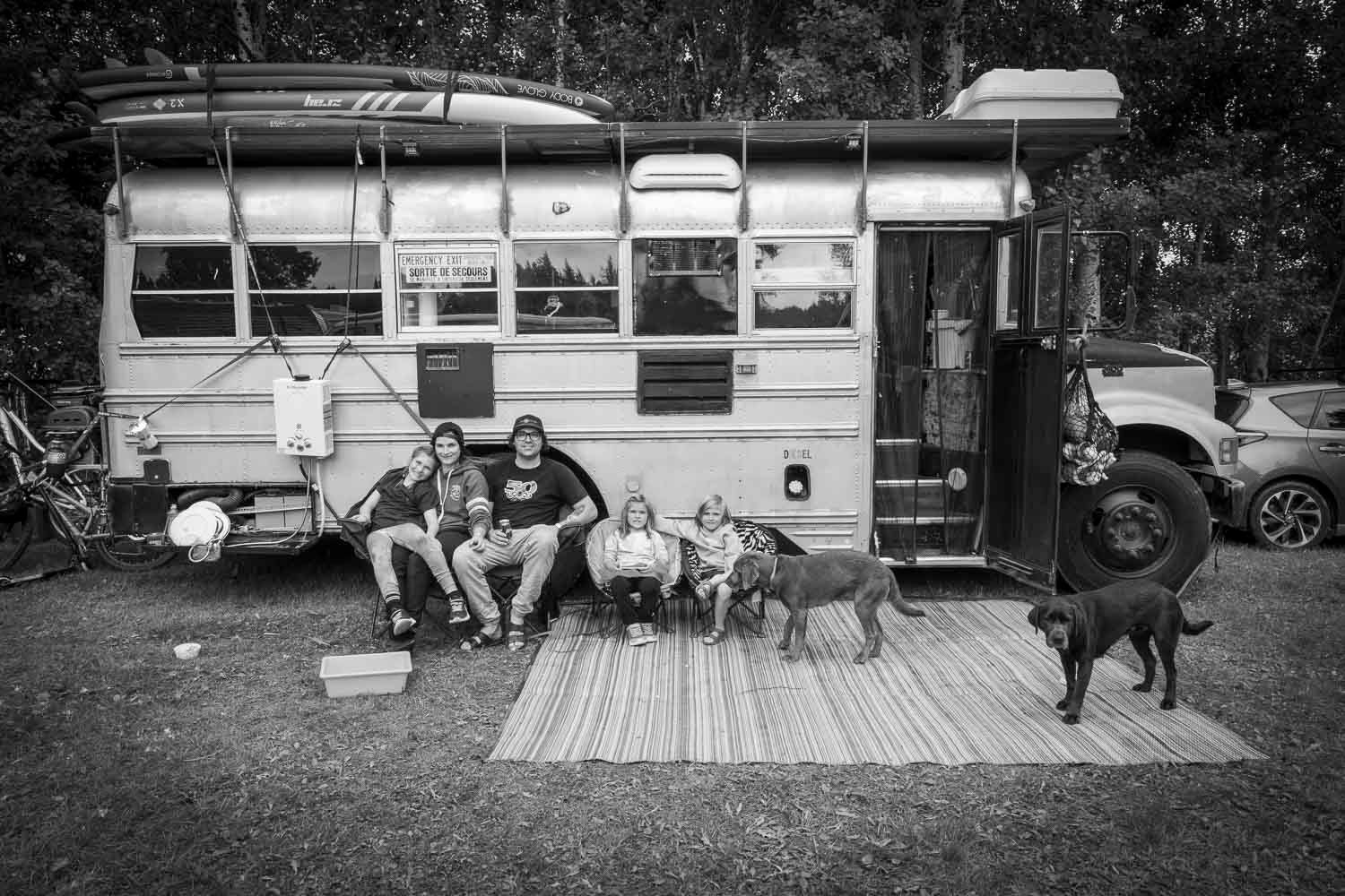 Famille, vanlife, autobus, chien,
Black and white photo of a family with dogs relaxing outside a converted school bus home on a striped rug.