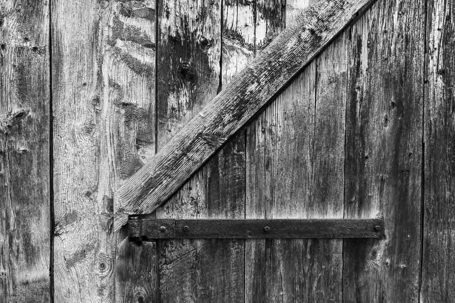 Black and white image of weathered wooden barn door with metal hinge.