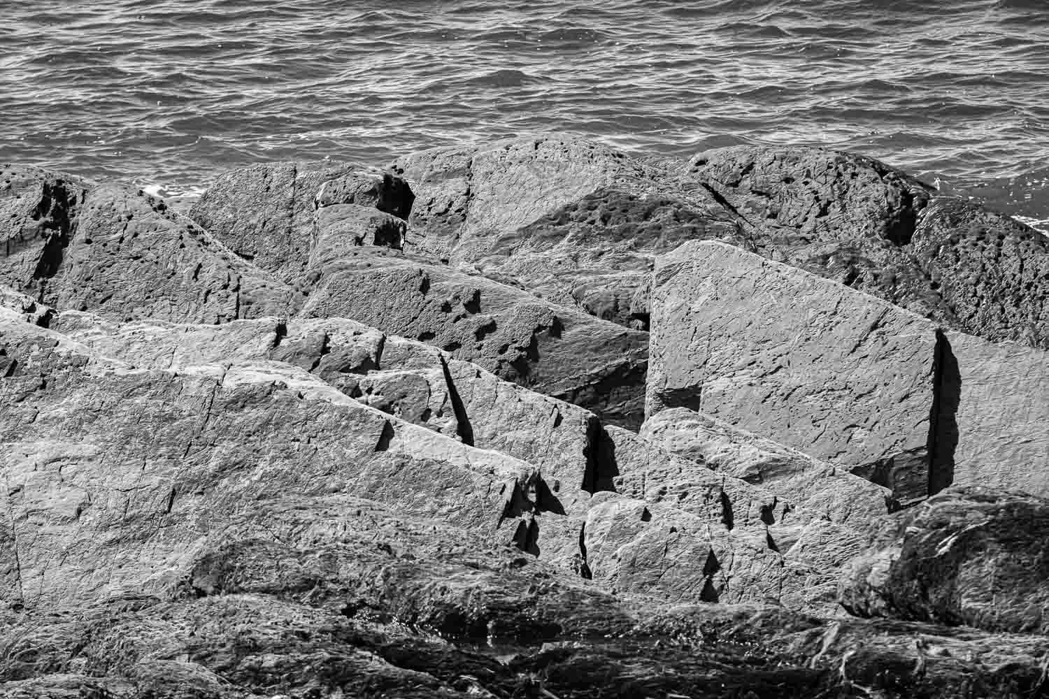 Black and white photo of large rocky breakwater boulders with ocean waves in the background.
