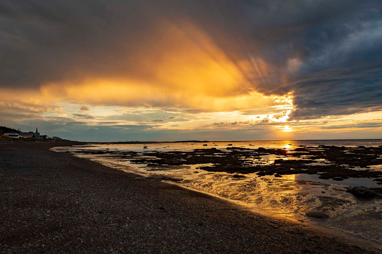Golden sunset over rocky coastline with dramatic storm clouds reflecting on wet sand and calm ocean waters.