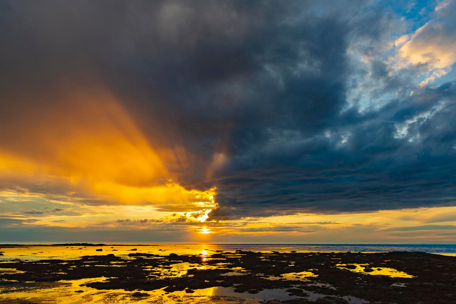 Coucher de soleil, Petit-
Dramatic sunset over ocean with golden light breaking through dark storm clouds reflecting on wet rocky shoreline.